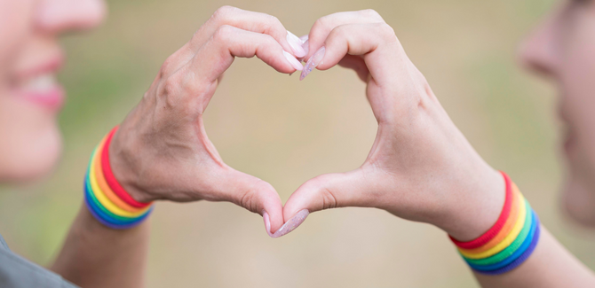 Two people with their hands formed together to make a heart. Both are wearing rainbow bands around their wrists.