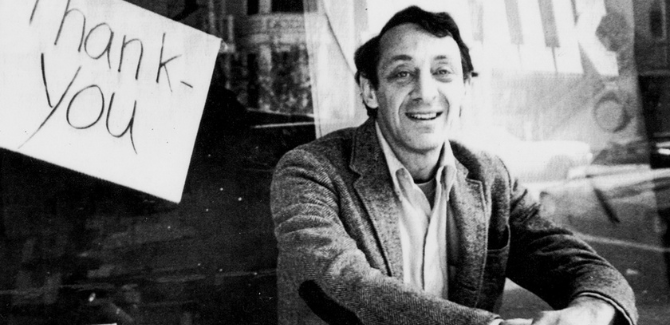 Harvey Milk sitting on steps wearing a suade jacked and shirt, campaign posters saying "MILK" and "Thank You" are behind him.