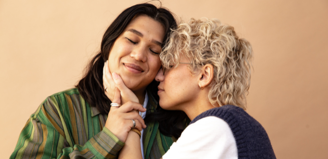 Two women, embracing each other in front of beige, orange background. Person on left has eyes closed, smiling, holding the hand of the other who is touching her face. Woman on right, has eyes closed, face is pressed to the side of other woman.