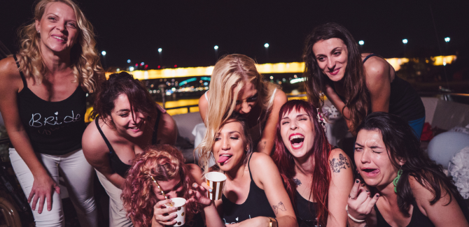 Group 8 of female friends wearing black shirts that say "bridal party". They are grouped together for a photo, but all are making silly (potentially drunk) faces. They appear to be having fun at a bachelorette party.