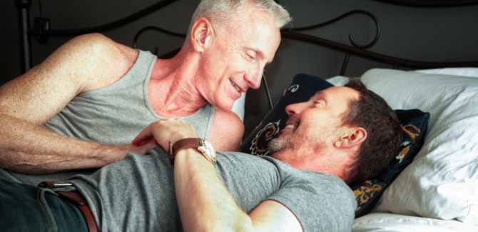 Two older men laying in bed, one on his back in gray t-shirt, the other man is wearing a gray tank and is on his side, intimately looking at the other man and holding his hand.