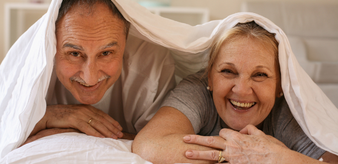 Older white couple, laying in bed together, on their stomachs facing the camera and smiling. Both are wearing gray t-shirts, and are huddled together under a sheet that is draped over their heads and bodies.