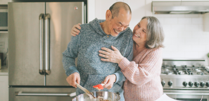Older couple, in a kitchen, standing over a mixing bowl, appearing to be baking. Man (LFET) is wearing a gray hooded sweatshirt, and is smiling with eyes closed. Woman (RIGHT) wearing pink sweater, left hand on his chest, smiling affectionately