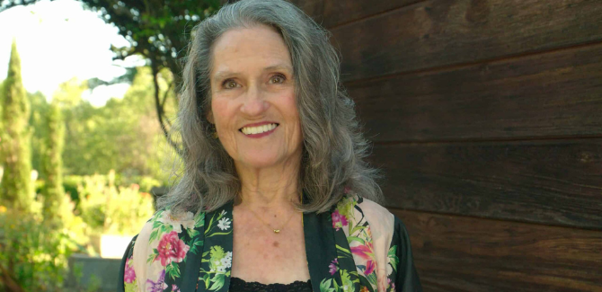 Joan Price, standing outside, wearing a floral top and hair long and wavy. She is smiling and standing just left of center in the frame.