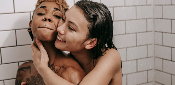 A Black woman and white woman in the shower together. White woman (behind) has their arms around Black woman in an embrace and is smiling. Black woman has eyes closed, head tilted up, and lips tightly pressed together in a focused pleasure.