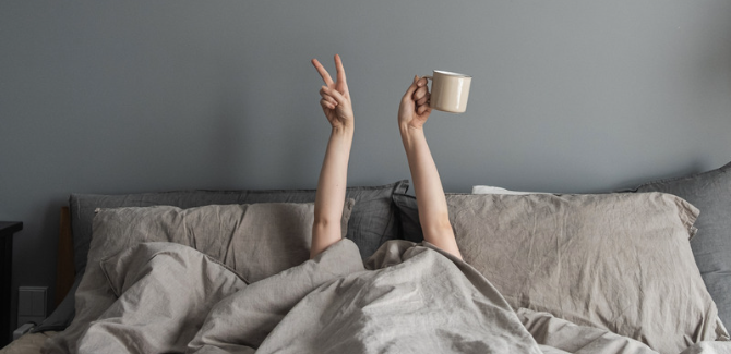 a grey/beige bed is shown with two pillows and sheets pulled over a person's face. Both arms are up and visible, the right hand with a peace sign, left with a coffee mug. The wall behind the person is grey. Person conveying contentness in bed.