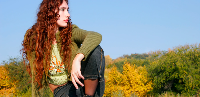 young person with long, red, curly/wavy hair, wearing jeans, green top and green sweater, sitting on the ground in the country. Their right knee bent upward and right arm resting on knee and they are looking a distance to the right.