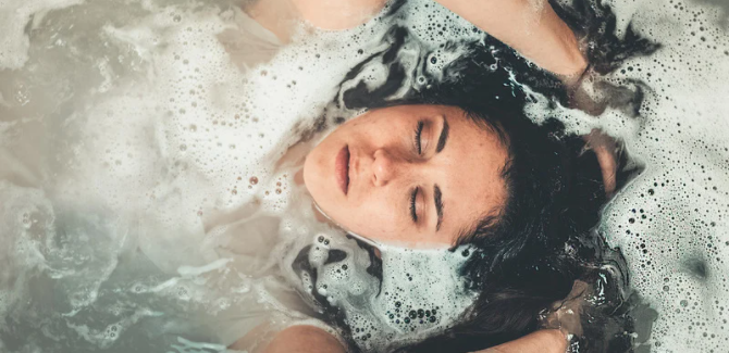 woman in bath tub. bubbles and water covering everything but face. Eyes are closed, hands over head, face relaxed