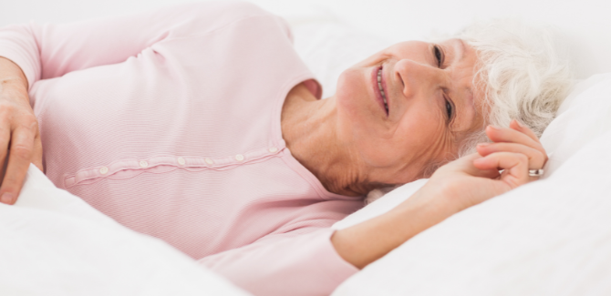 Older woman laying in bed on her side, wearing blush pajama top, slightly smiling.