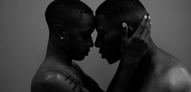 Two black men, standing together with foreheads pressed together in loving, intimate embrace. Man (left) has their hands around partner's (right) head. Both men are shirtless and frame shows from shoulders up.