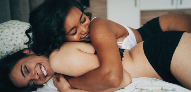 Image of two women laying together, one on top of the other, with her arms around her. Both are smiling and wearing bras and panties.