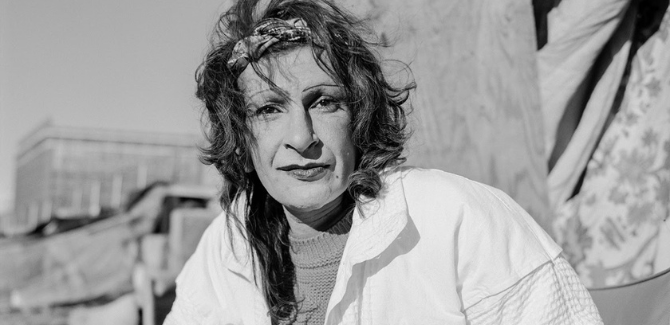 Sylvia Rivera standing in a street, looking at the camera with straight face, wearing a white jacket, hair is tossled, and floral head band. Image is in black and white.