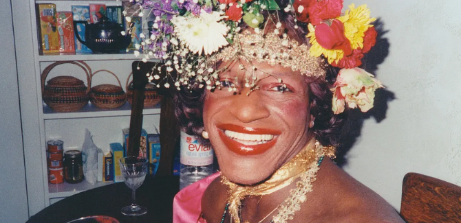 Marsha P Johnson sitting at a table inside house, wearing colorful flowers in their hair, pink dress, gold necklaces and ribbons around their neck, smiling. Marsha has a bold red lip, pink rouge on their cheeks and is smiling.