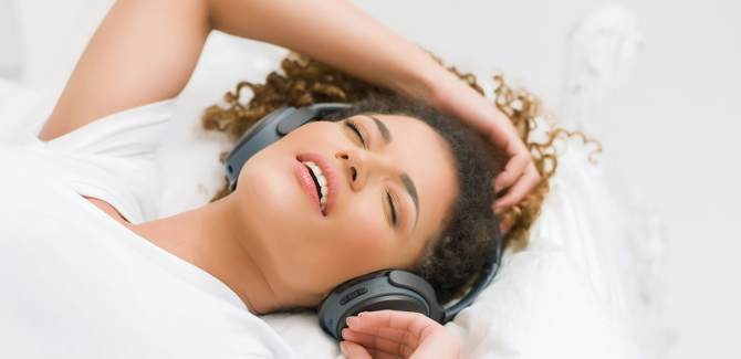 black woman laying in bed with white sheets and pillows, their right arm over head, listening to music on large black headphones. Their eyes are closed and small grin on their face.