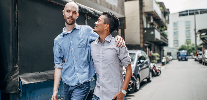 Interracial gay couple walking together on a sidewalk. Man (LEFT) is all, bald, white, and arm around partner's shoulder. Man (RIGHT) shorter, asian, with arm around partner's waist, smiling at him.