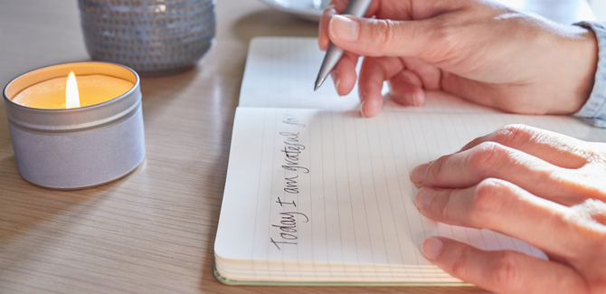 White hands writing in a journal "Today I am grateful for" with a candle burning next to the journal