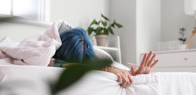 Person with blue hair, face down on bed, grasping at sheets and edge of bed in pleasure.