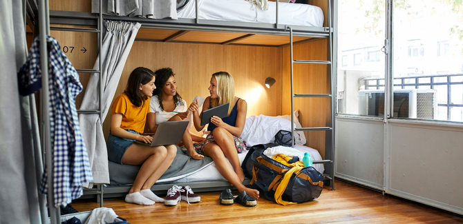 Diverse college females sitting on bottom bunk of bed, talking to each other, and pointing at computers and tablets to show a studious discussion.  