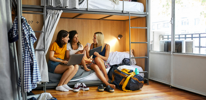 Diverse college females sitting on bottom bunk of bed, talking to each other, and pointing at computers and tablets to show a studious discussion.