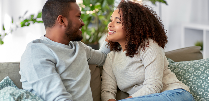 Black couple on the couch, talking, smiling at each other. Man (LEFT) wearing gray crew sweatshirt, woman (RIGHT) wearing brown long sleeve shirt with jeans.