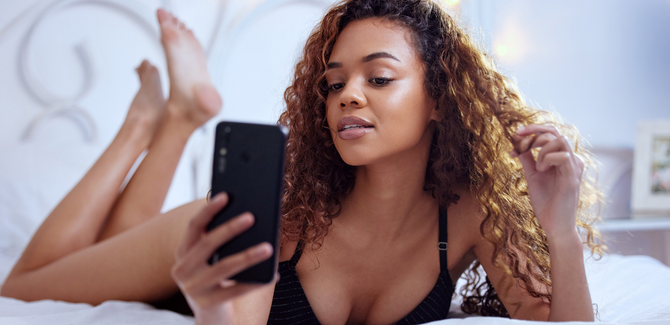 Black woman, laying on bed in black undergarments, legs kicked up behind her, posing sexy for her phone.