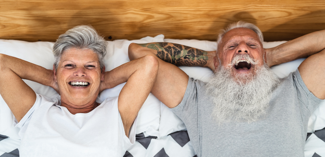 Older white couple, laying in bed face up, side-by-side. Arms are placed behind their heads and both are looking up and smiling wide. Woman (left) has short gray hair and white tee. Man (RIGHT) has gray hair, full gray beard, and gray shirt.