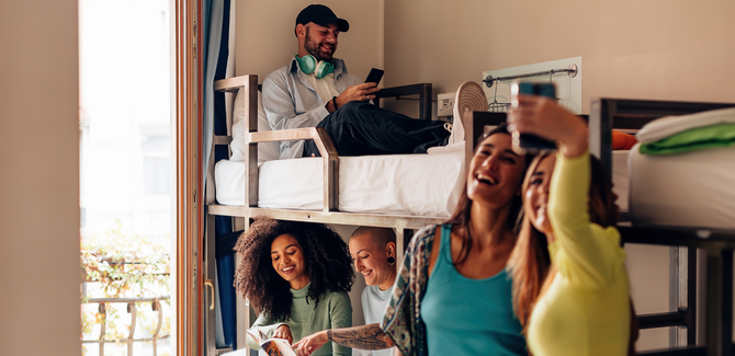 Diverse students hanging out in a dorm room. In foreground, two female students are taking a selfie, background (bottom bunk) two students are studying together, and top bunk, man is listening to music on phone