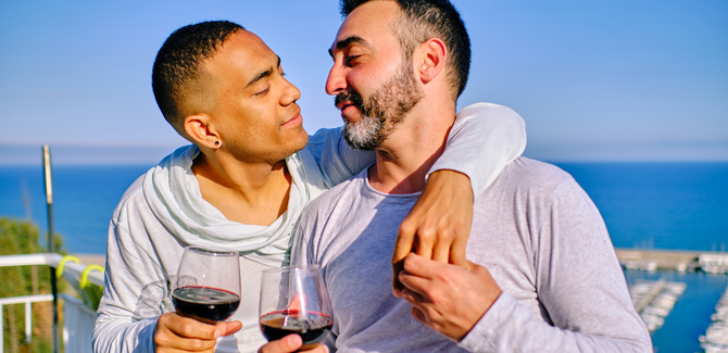 Black man and white man standing near a boating harbor with glasses of red wine and are lovingly looking into each other's eyes. Black man (left) has his left arm wrapped around partner's neck and shoulder, white man (right) is holding his hand.