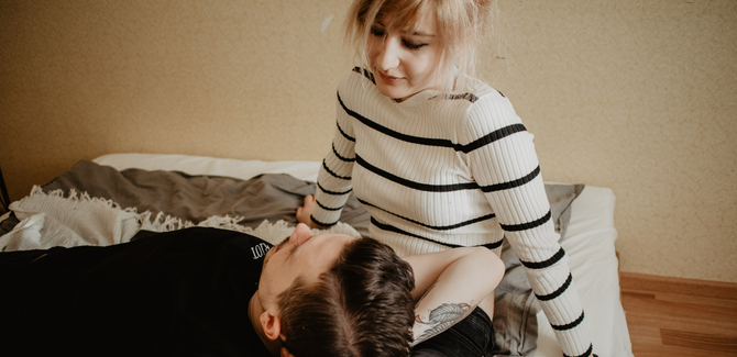 White couple, on bed, talking. Man (laying down on partner's lap) is wearing black shirt, and has tattoos. Woman (sitting on bed, looking down) is wearing white and black striped sweater.