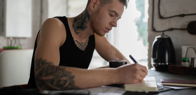 Tattooed white man journaling at a kitchen counter while drinking coffee