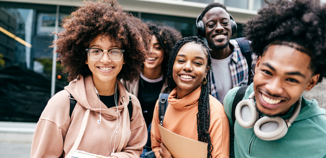 Group of black students on campus in colorful sweatshirts with backpacks, books, all smiling at the camera in a clustered and organic manner.