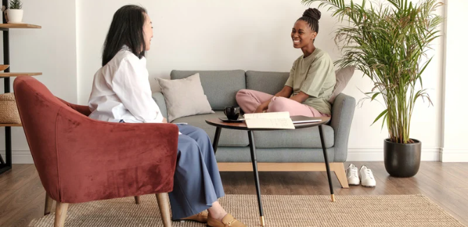 [left] woman sitting in burnt orange/maroon chair with blue pants and white top, facing another woman opposite her [right] on a grey couch with pink pants and green/grey top. Both are smiling, appearing to be in session.