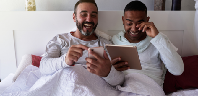 couple in bed, sitting up, laughing and smiling. Man on left is in long-sleeved gray shirt, on his phone. Man on right is also wearing a long-sleeved gray shirt wiping eye, laughing, holding a tablet.