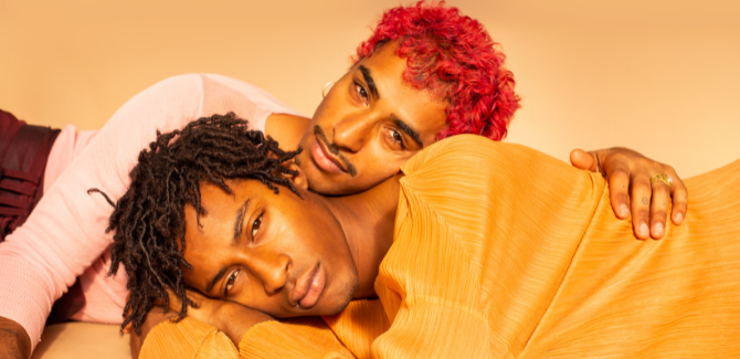 Two queer individuals, laying on the ground, in front of orange backfround. Person in front is laying on their right side, while person behind, lays on left side, their head laying on the other's neck, and left hand holding the other's side.