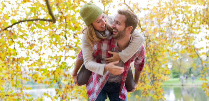 Man in red flannel shirt carrying a woman in grey sweater and green beanie hat on his back, playfully. They are outside, under a fall tree.