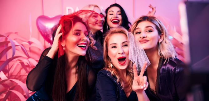 Five girls huddled together taking a group picture with smiles and silly faces. They are wearing all black and have a pink hue to the lighting to portray a bachelorette party.