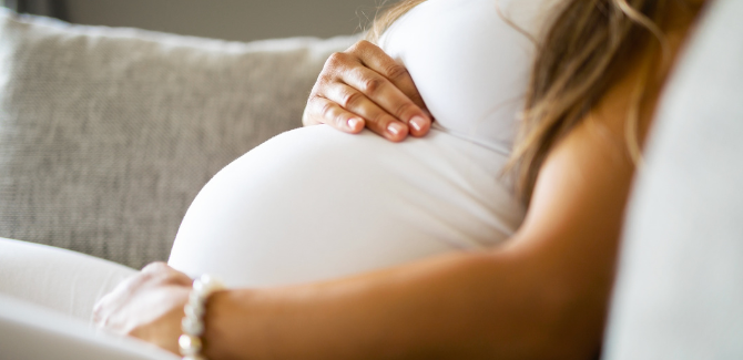 Image of a pregnant person sitting on a couch, wearing white shirt, right hand on top of stomach and left hand on bottom of stomach to show baby bump.