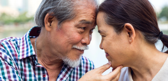 Senior asian man and woman intimately close with their foreheads pressed together as they smile and gaze at one another. The man has his right hand on her chin.