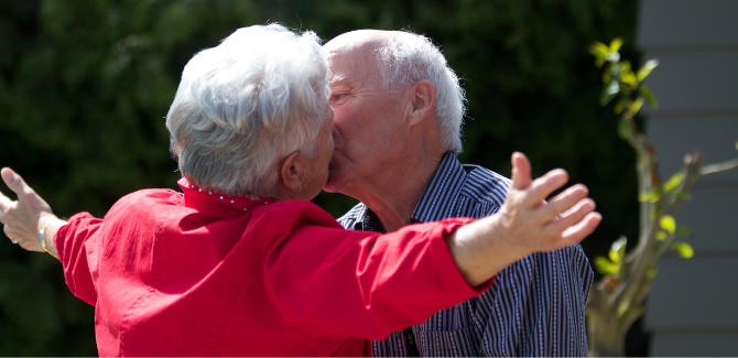 Older white couple, standing outside, kissing. (LEFT) Person wearing coral, long-sleeved top and gray hair has their arms stretched out as man (RIGHT) wearing blue striped shirt and gray hair kisses them. His eyes are slightly open.