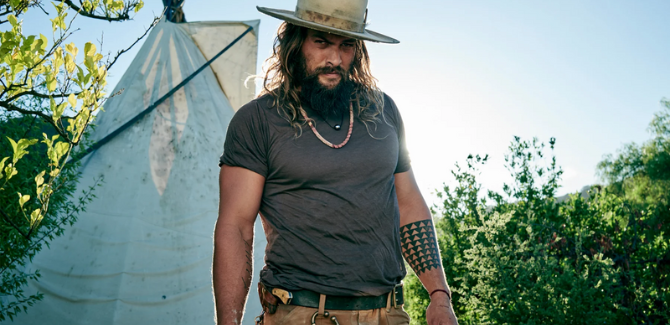 Jason Momoa standing outside next to a tent wearing gray top, khakis, and white hat.