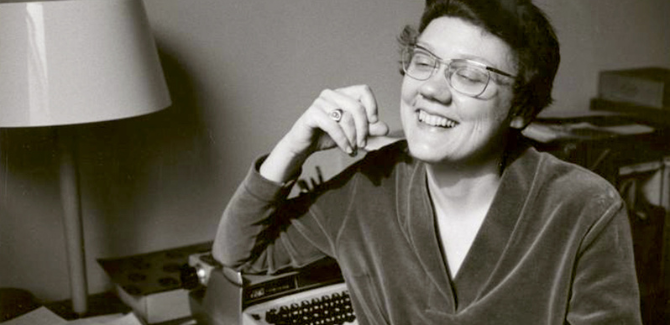 Barbara Gittings sitting at a desk, next to a type righter, wearing a velvet-like sweater, smiling, looking off-camera. Image is in black and white.