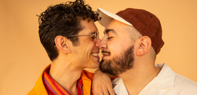 Two men, facing each other, about to kiss, both are smiling in front of an orange background.