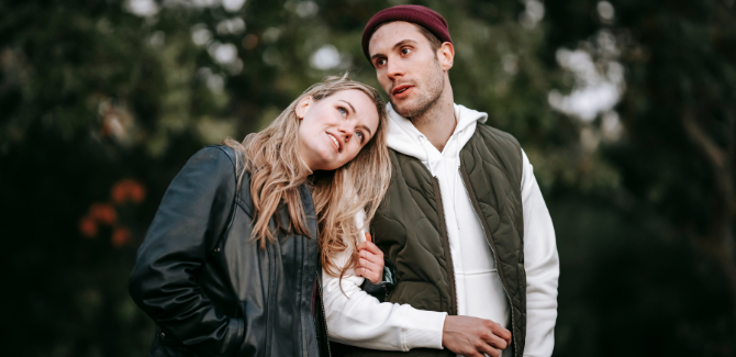 Couple walking outside in a wooded area. Woman is on left, wearing black leather jacket, with her head resting on mans shoulder and her left arm linked with his. He is wearing a white hoodie, brown fest, and red beanie.