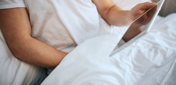 person sitting in bed in white t-shirt, holding a tablet in left hand, with right hand down their pants, but it is covered by the blanket to insinuate masturbation
