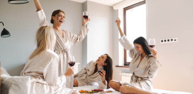 Four girls sitting on a bed wearing white robes drinking red wine and eating a charcuterie board of meats, cheeses, and small foods. They are celebrating and cheering.