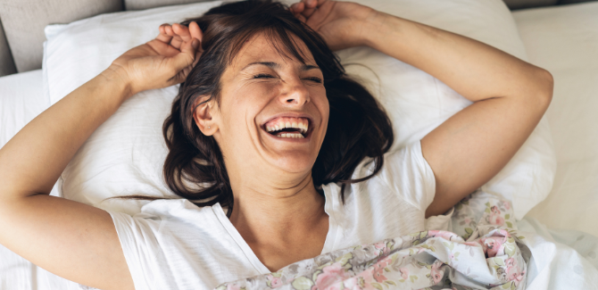 Woman laying bed, wearing a white tshirt, smiling, with hands over head, and eyes closed.