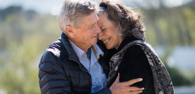 Older couple sitting outside, embracing in a side hug. Woman is smiling with her head pressed into the man's shoulder as he embraces her.