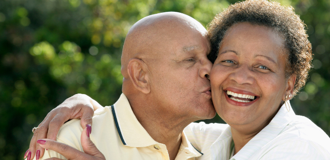 Older black couple outside posing for picture, embracing each other. Man (LEFT) is kissing the cheek of (RIGHT) woman and her arms are wrapped around his shoulders and chest in a side, hug.