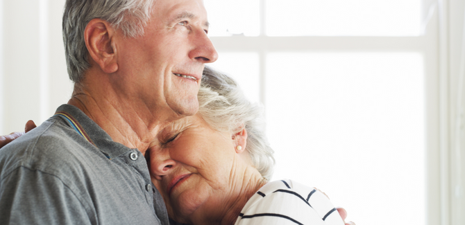 Older couple standing together, intimate embrace. Man is facing to the right, staring ahead, smiling. Woman is standing slightly in front of him, with her head on his left shoulder, eyes closed, and right hand wrapped around his shoulder blades. 