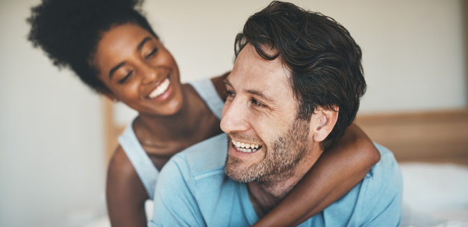 Mixed race couple in bed playing together. Black woman is kneeling behind white man with her left arm wrapped around his shoulders and chest. He looks back at her and both are smiling.